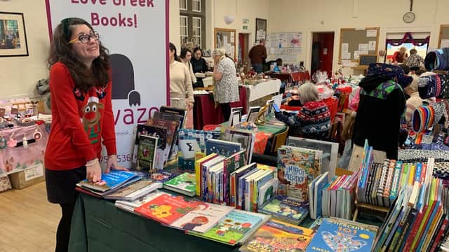 A woman in a red sweater smiles at a table filled with colorful Greek books at a bustling event in Sheffield.