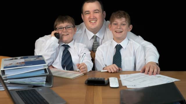 A man and two boys in white shirts and ties sit at a desk with paperwork and a laptop, smiling at the camera.
