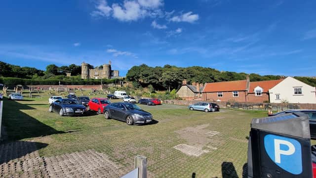 St Mary's pay and display car park with parked cars, green grass, and a historic building on a hill under a blue sky.
