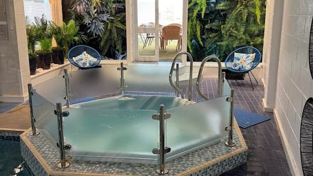 Hexagonal hot tub surrounded by lush greenery, with blue chairs and a doorway leading to a bright outdoor area.