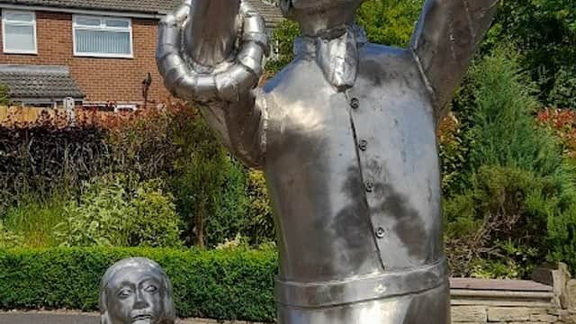 Metal sculptures of a man holding a shovel and a girl, set in a green park with residential buildings in the background.