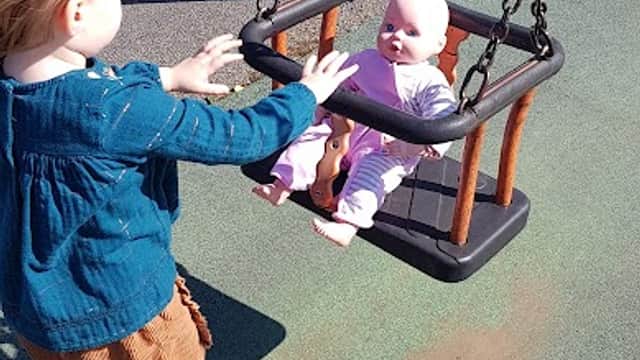 A child pushes a doll in a swing at a playground with colorful surfaces and a clear blue sky in Wombwell Skate Park.