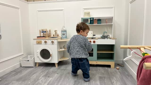 A toddler playing in a miniature kitchen with a toy stove, sink, and washing machine in a bright, playful room.