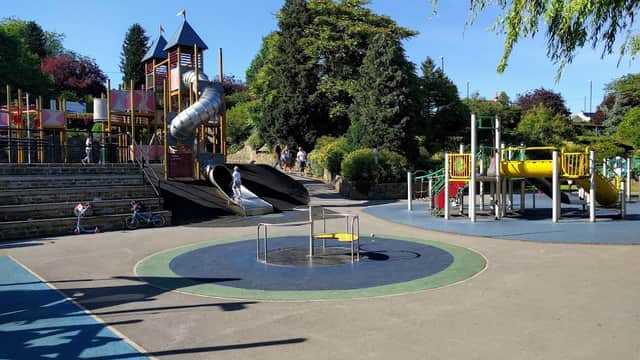 Playground featuring slides, climbing structures, and a spinning carousel, surrounded by greenery and a clear blue sky.