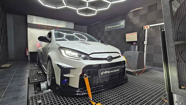 White Toyota car on a dynamometer in a high-tech performance testing facility with hexagonal ceiling lights.