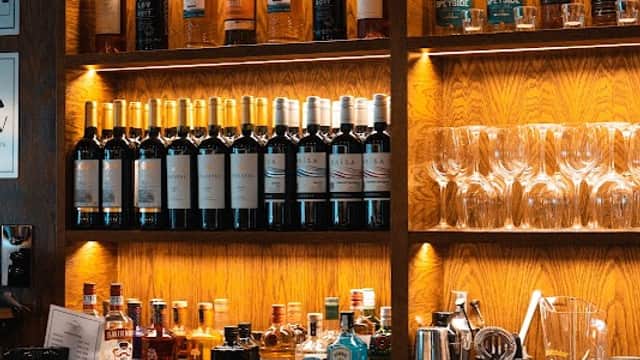 Wooden bar with shelves displaying various bottles of spirits and glasses, illuminated by warm lighting.