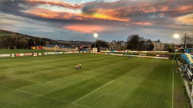 Football pitch at Stocksbridge Park Steels FC under a colorful sunset, with players warming up and floodlights on.