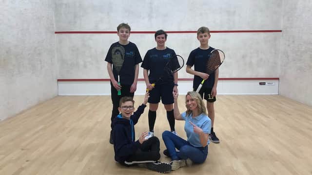 Four young squash players and a coach pose on a squash court, holding racquets and smiling at the camera.