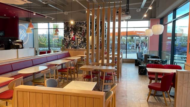 Modern coffee shop interior with wooden furniture, red seating, and large windows showing a drive-thru sign outside.