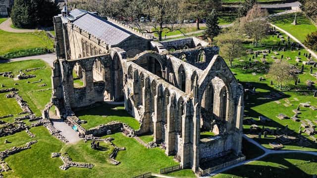 Ruins of Bolton Abbey with arched stone walls, surrounded by green grass and trees, near a cemetery.