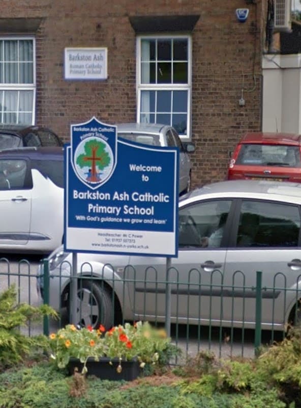 Welcome sign for Barkston Ash Catholic Primary School with colorful flowers and parked cars in front.