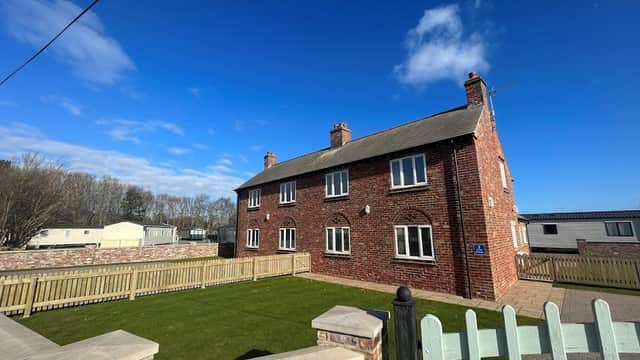 Red brick building with multiple windows, surrounded by a green lawn and wooden fence under a clear blue sky.