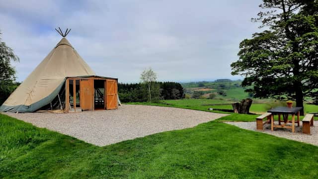 A large teepee tent on a gravel area, surrounded by lush green grass and trees, with rolling hills in the background.