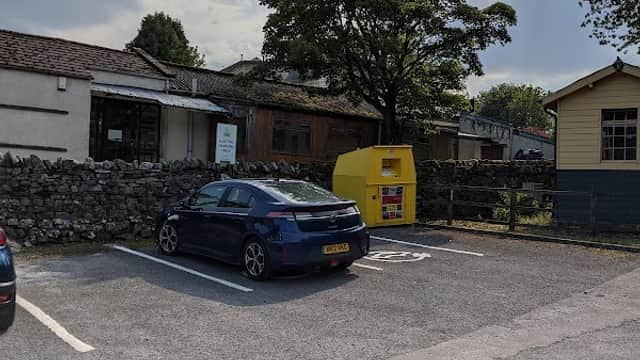 Pod Point charging station beside a parked car, with a stone wall and trees in the background in Burtersett, Yorkshire.