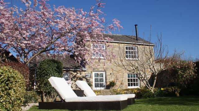 Charming stone cottage with blooming cherry trees and sun loungers in a lush garden, set against a clear blue sky.