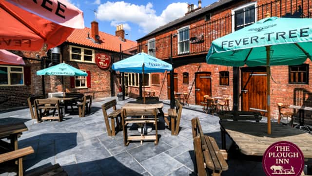 Outdoor seating area with wooden tables and colorful umbrellas at The Plough Inn, surrounded by brick buildings.
