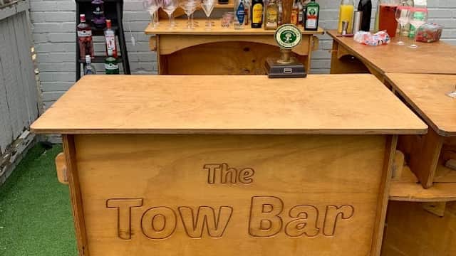 Wooden bar setup with "The Tow Bar" sign, shelves of drinks, and decorative flowers in a green outdoor space.