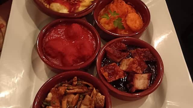 Five small bowls of colorful tapas dishes on a white platter, featuring various sauces and toppings.