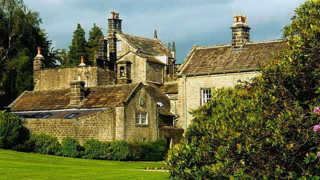 Historic stone building with multiple chimneys, surrounded by lush greenery and blooming shrubs under a cloudy sky.