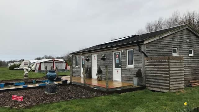 Wooden lodge with two doors, garden area, and a fountain, surrounded by grassy fields and a tent in the background.