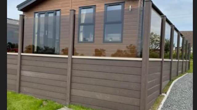 Modern holiday cabin with large windows, surrounded by a wooden fence and green grass, set against a cloudy sky.