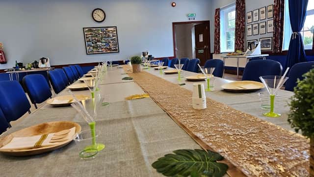 Long table set with green and gold decor, plates, glasses, and a clock on the wall in a bright, airy hall.