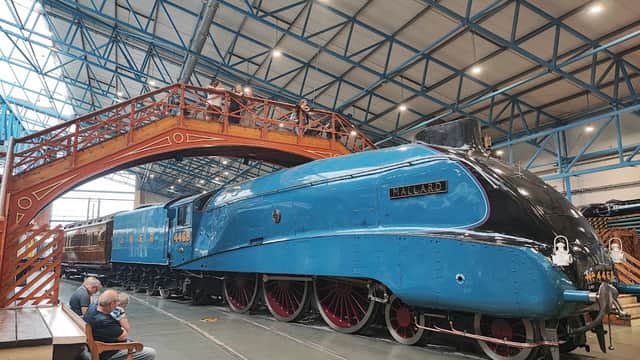 Blue steam locomotive "Mallard" displayed indoors beneath a high ceiling, with wooden bridge and visitors nearby.