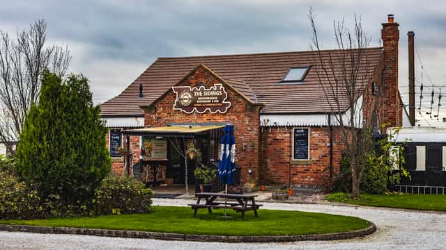 Red-brick building with a sign reading "The Sidings", outdoor seating, and greenery in a rural setting.