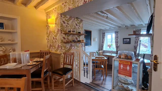 Cozy tea room with wooden furniture, floral wallpaper, and a display of baked goods near a sunny window.