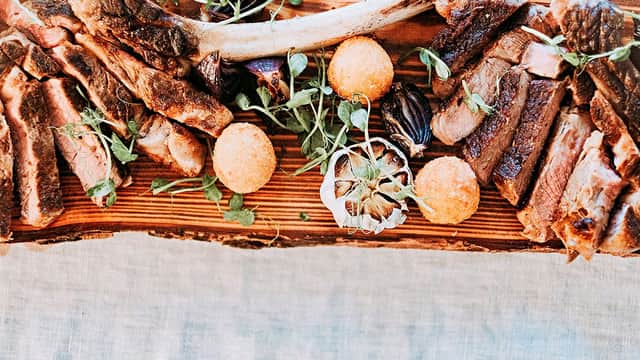 A wooden platter featuring an assortment of grilled meats, garnished with herbs and round orange croquettes.