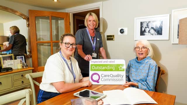 Two caregivers and an elderly woman holding an "Outstanding" Care Quality Commission sign at a cozy home setting.