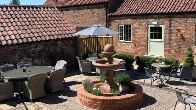 Outdoor seating area with a fountain, surrounded by brick walls and buildings under a clear blue sky.