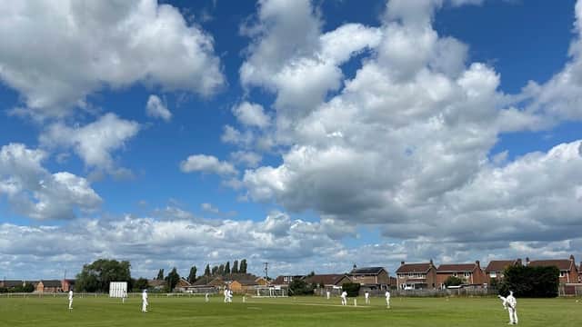 Cricket match in progress on a green field under a blue sky with fluffy clouds, surrounded by houses.