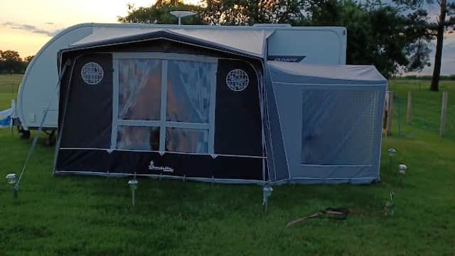 Caravan with a black awning set on green grass, surrounded by trees and a colorful sunset sky.