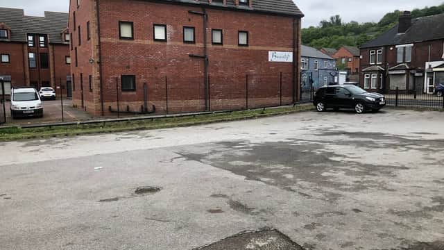 Free parking area with a gravel surface, surrounded by brick buildings and a few parked cars. Overcast sky.