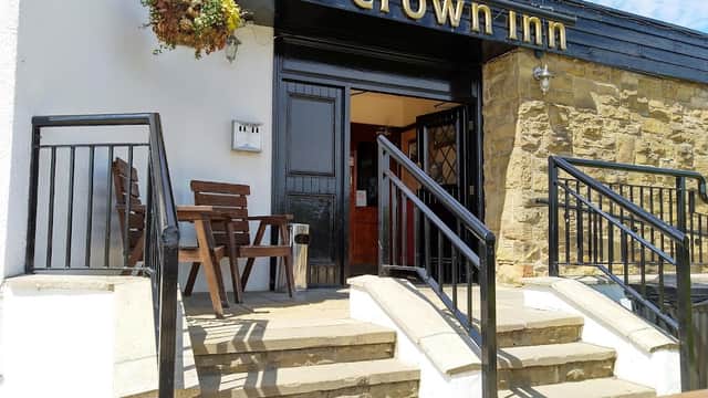 The Crown Inn entrance with wooden chairs, stone steps, and a hanging flower basket under a blue sky.