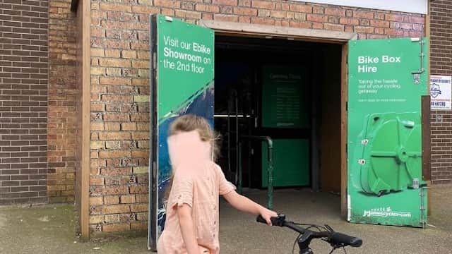 Child with a bike stands outside J E James Cycles shop, featuring a green sign and a bike hire display.