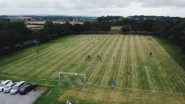 A green football pitch with players and spectators, surrounded by trees and parked cars under a cloudy sky.