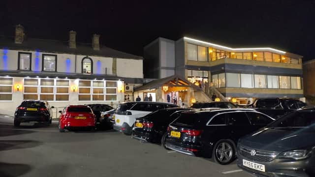 Car park at night with several parked cars, a modern building with bright lights, and a traditional structure nearby.