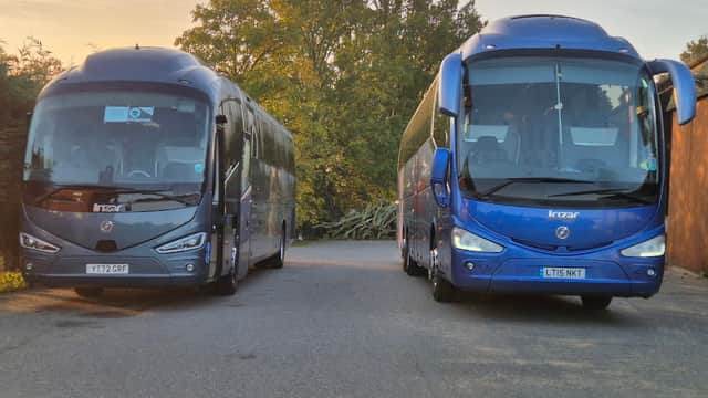 Two blue coaches parked side by side on a road, surrounded by trees in the background, during sunset.