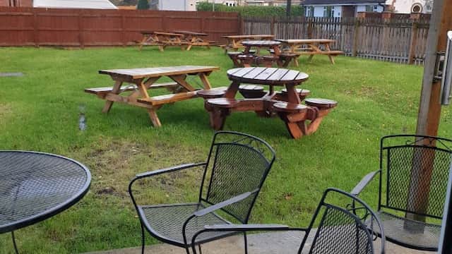 Outdoor seating area with picnic tables on green grass, surrounded by wooden fencing and residential buildings in the background.