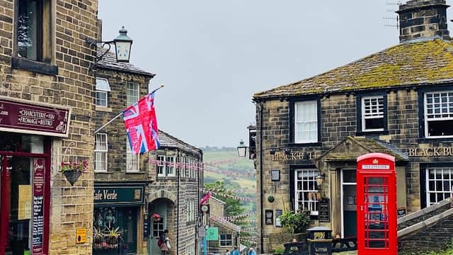 Quaint cobblestone street with shops, a red telephone box, and flags, surrounded by green hills under a cloudy sky.