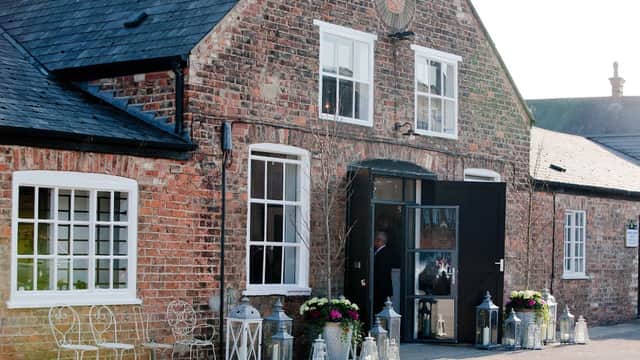 Charming brick building with large windows, black door, and flower arrangements beside lanterns and outdoor seating.