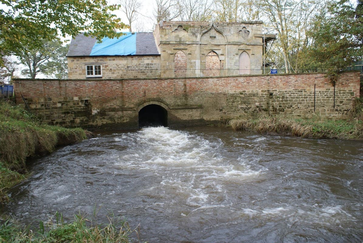 Stone building with arched entrance beside a flowing river, surrounded by trees and autumn foliage.