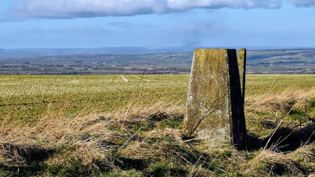 Trig point stands in a grassy field, overlooking a vast landscape with rolling hills under a partly cloudy sky.