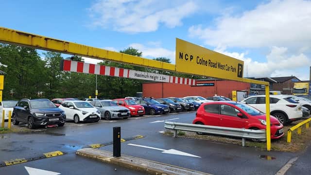 NCP Colne Road car park with parked cars, yellow signage, and trees in the background under a cloudy sky.