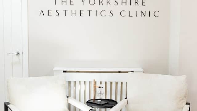 Two stylish white armchairs with a small black table between them, featuring the Yorkshire Aesthetics Clinic logo on the wall.