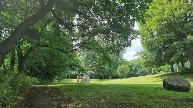 Lush green park with large trees, a grassy area, and stone sculptures under a partly cloudy sky.