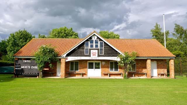 Scruton Cricket Club building with a red-tiled roof, green lawn, and cloudy sky, featuring a sign and scoreboard.