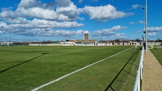 Green football pitch with white lines, a blue sky, and clouds; background features a building and a tower.